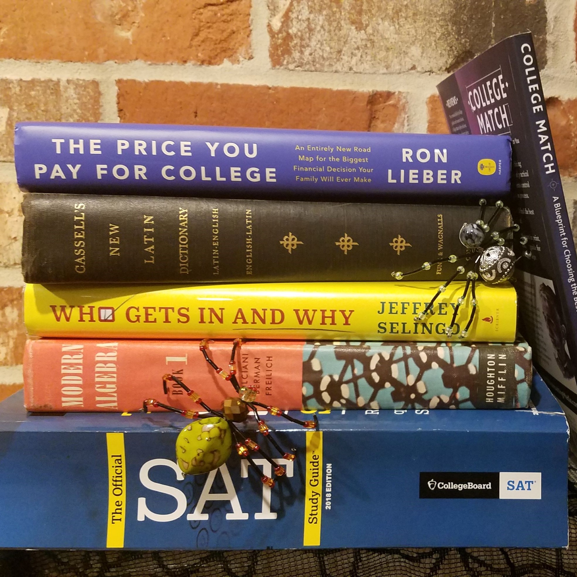 A stack of books, including The Price You Pay for College, Who Gets In and Why, College Match, an SAT study guide, a Latin dictionary, and a vintage algebra book. Two beaded spiders are positioned crawling on the books.