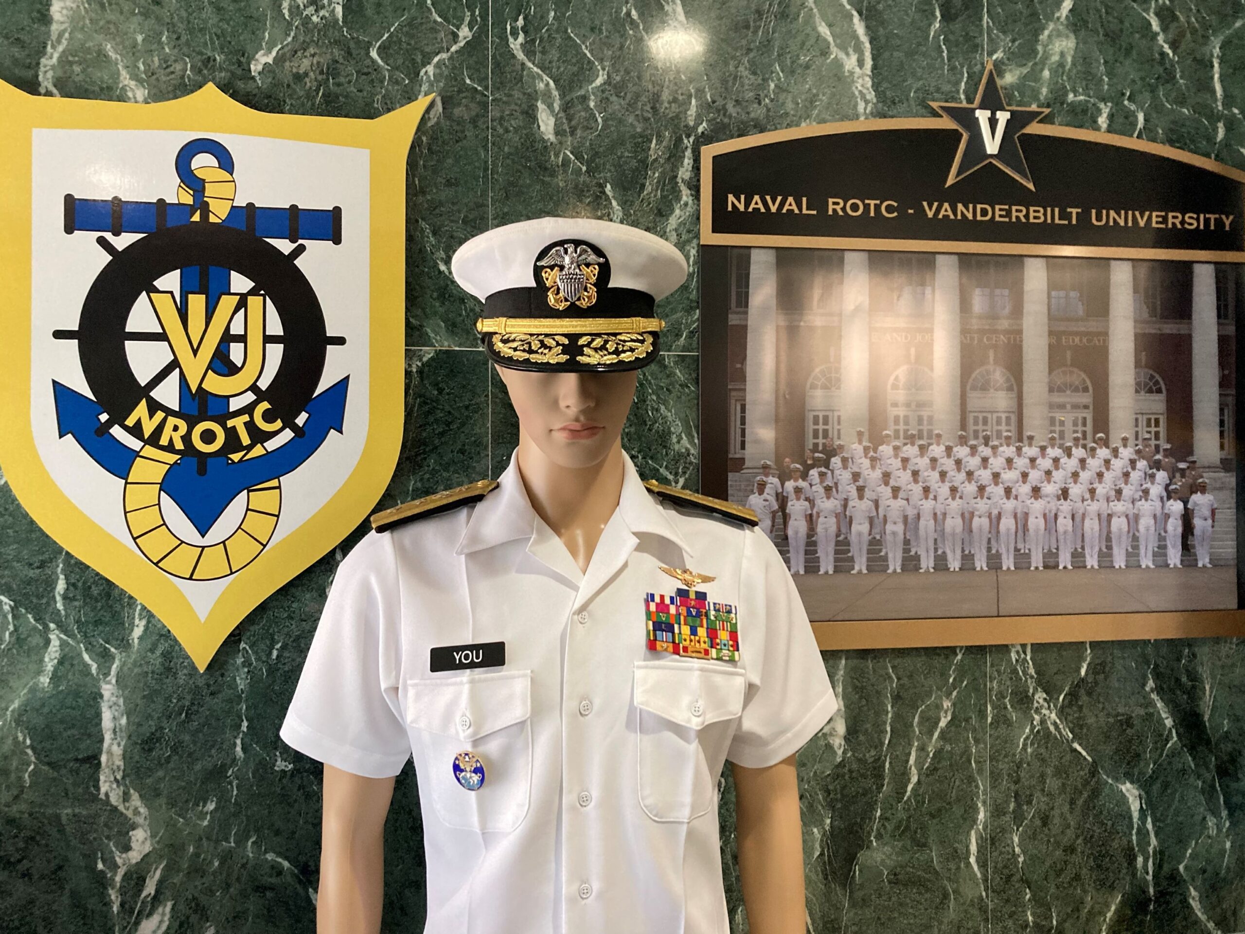 Display at Vanderbilt Navy ROTC unit, including the unit crest, a photo of current midshipmen, and a mannequin wearing a summer white uniform with two star admiral insignia, aviator wings, and a nametag for "YOU"